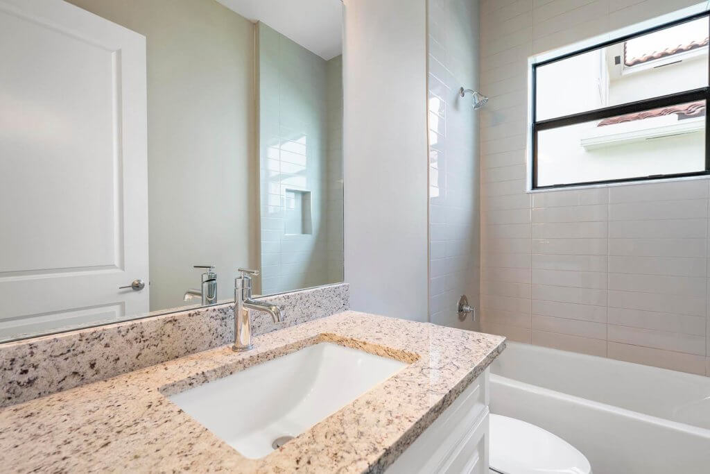 Modern bathroom at Villamar at Toscana Isles featuring a granite countertop with a single sink and chrome faucet, a bathtub with showerhead, a large mirror, and a window allowing natural light. Walls are painted in light hues, harmoniously complementing the tiled area around the tub.