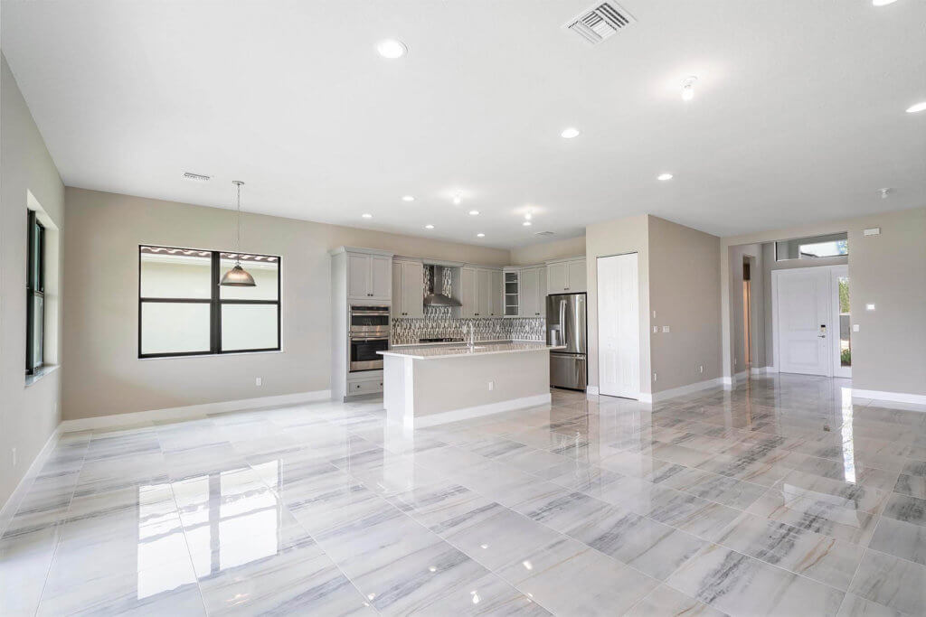 Spacious modern kitchen and living area at Villamar at Toscana Isles with white walls and glossy marble tile flooring. The kitchen features a large island, stainless steel appliances, and ample cabinets. Multiple recessed lights illuminate the space, while large windows offer natural light.