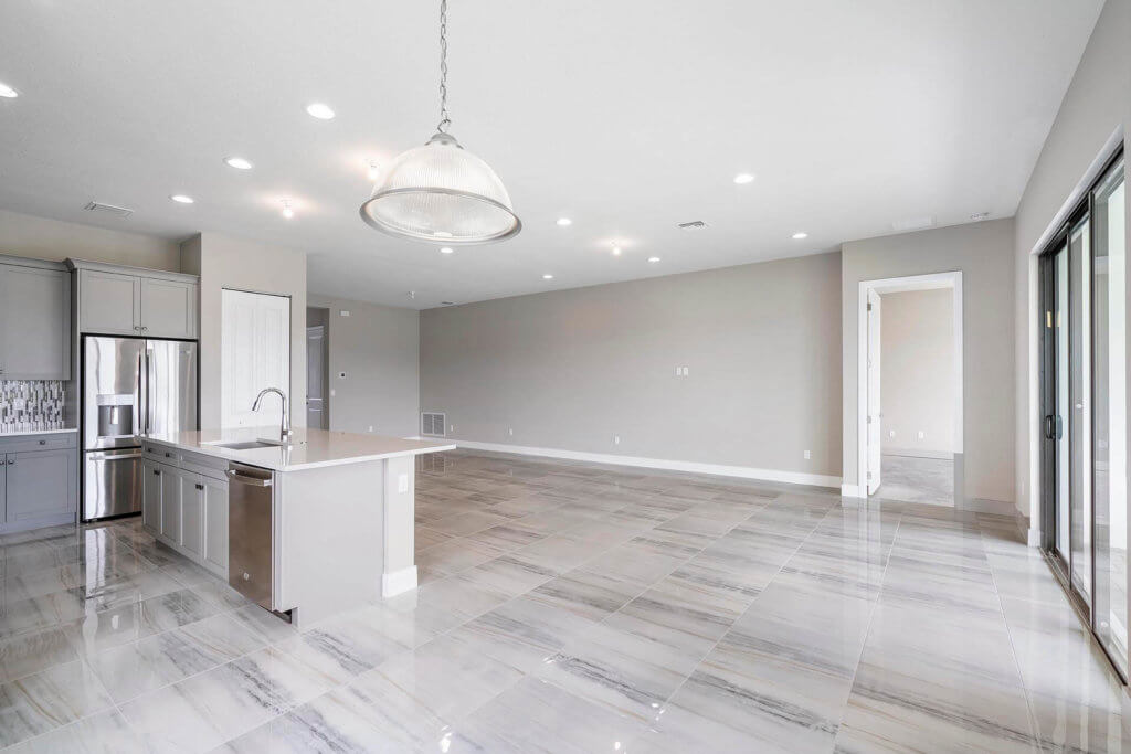 The spacious, modern kitchen and living area at Villamar at Toscana Isles boasts light gray tiled floors, a center island with a sink, stainless steel appliances, and elegant light gray cabinets. Sliding glass doors invite you to explore the next room.