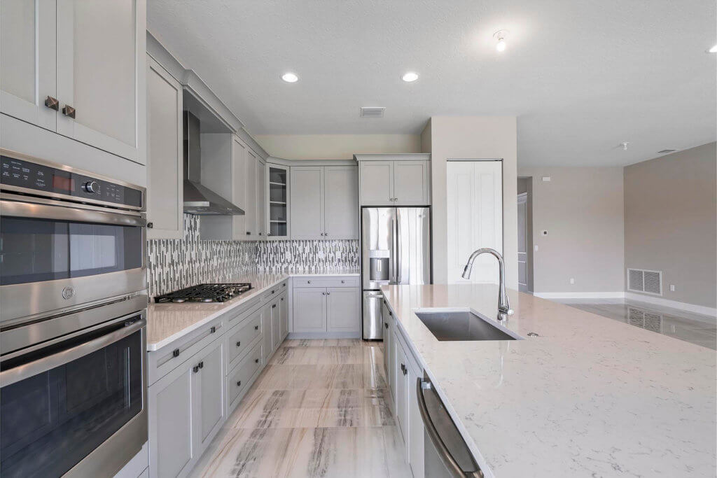 A modern kitchen at Villamar at Toscana Isles showcases marble countertops, light gray cabinets, and stainless steel appliances, with a double oven and refrigerator. The metallic-patterned backsplash and light wood-finish tiled floor add an elegant touch to the space.