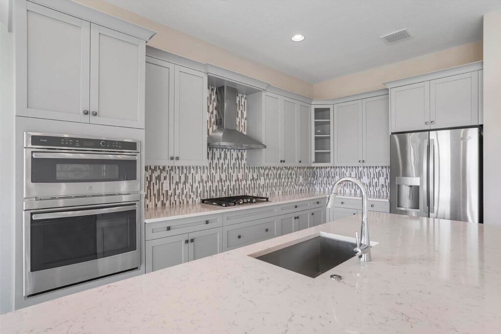 Experience the elegance of a modern kitchen at Villamar at Toscana Isles, featuring light gray cabinets, a stainless steel stove and oven, and a sleek silver vent hood. A mosaic tile backsplash complements the large white quartz countertop with an integrated sink, while a stainless steel refrigerator stands on the right.