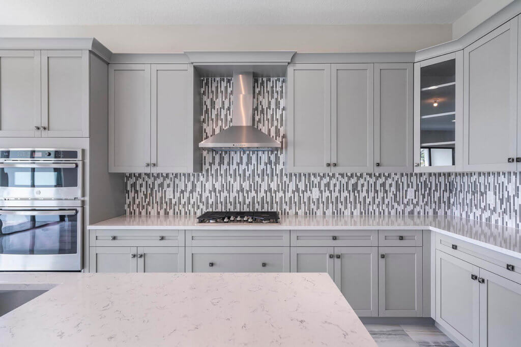 A modern kitchen at Villamar at Toscana Isles features gray cabinets and a white marble island. The backsplash displays a vertical pattern of gray, white, and black tiles. Stainless steel appliances, including a range hood and double ovens, are seamlessly integrated into the design.