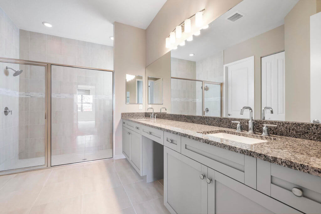 The spacious bathroom at Villamar at Toscana Isles features a large granite double sink vanity with silver faucets, a glass-enclosed shower, light beige walls, and tiled floors. Three lights hang above the mirror, and there's a closed door in the corner.