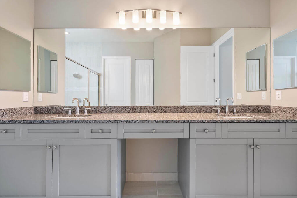 A modern bathroom at Villamar at Toscana Isles features a large mirror, dual sinks with granite countertops, gray cabinetry, and overhead lighting. A glimpse of a shower door and white door adds depth to the reflection.