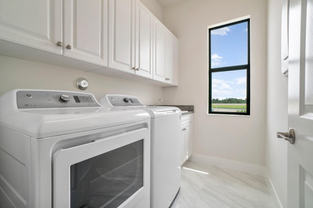 In the Villamar at Toscana Isles community, a bright laundry room features a white washing machine and dryer. White cabinets line the walls above, while a countertop with a sink is visible. A window lets in natural light, offering a serene view of greenery outside.