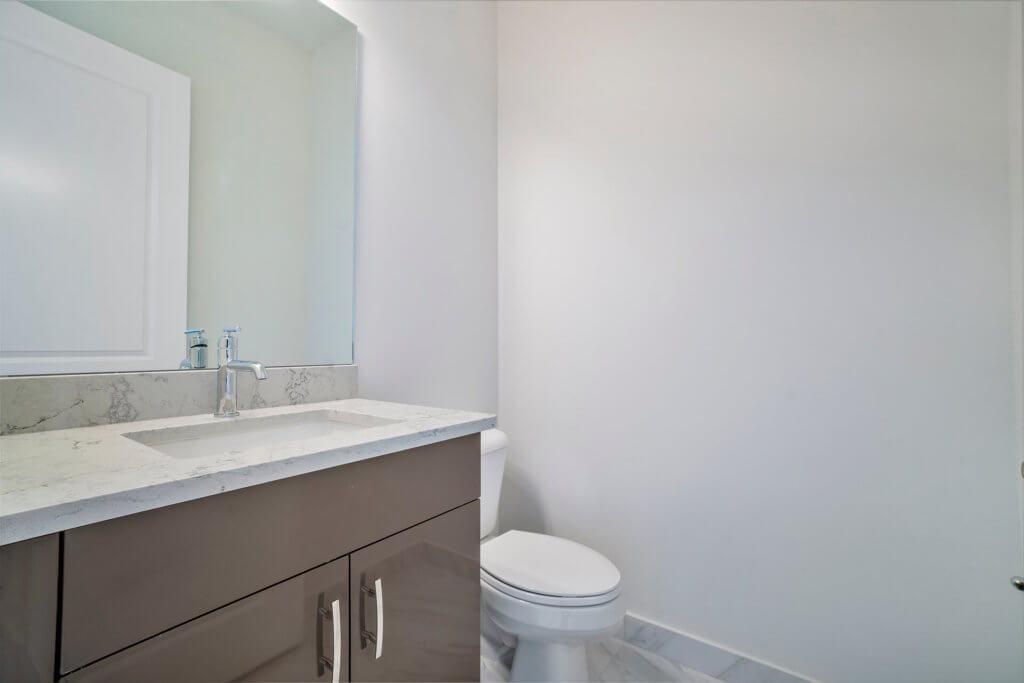 A modern bathroom at Villamar at Toscana Isles boasts a sink with a marble countertop and sleek faucet. A large mirror hangs above, reflecting elegance. To the right, a toilet complements the plain, light-colored wall, while tiled floors enhance the contemporary vibe.