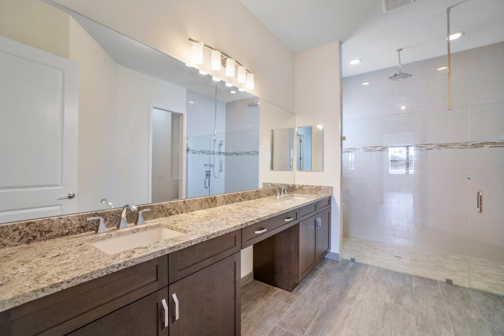 The modern bathroom at Villamar at Toscana Isles boasts a long granite countertop with dual sinks and dark wood cabinets. A large mirror is elegantly mounted above, while the spacious glass-enclosed shower area features soothing beige tiles.