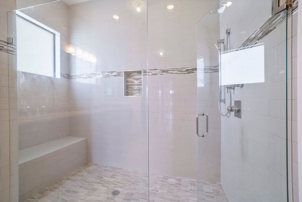 A spacious shower area at Villamar at Toscana Isles features white tiled walls and a glass door. Inside, a tiled bench sits to the left, with multiple showerheads on the right. A window and decorative tile strip enhance natural light and detail in the serene space.