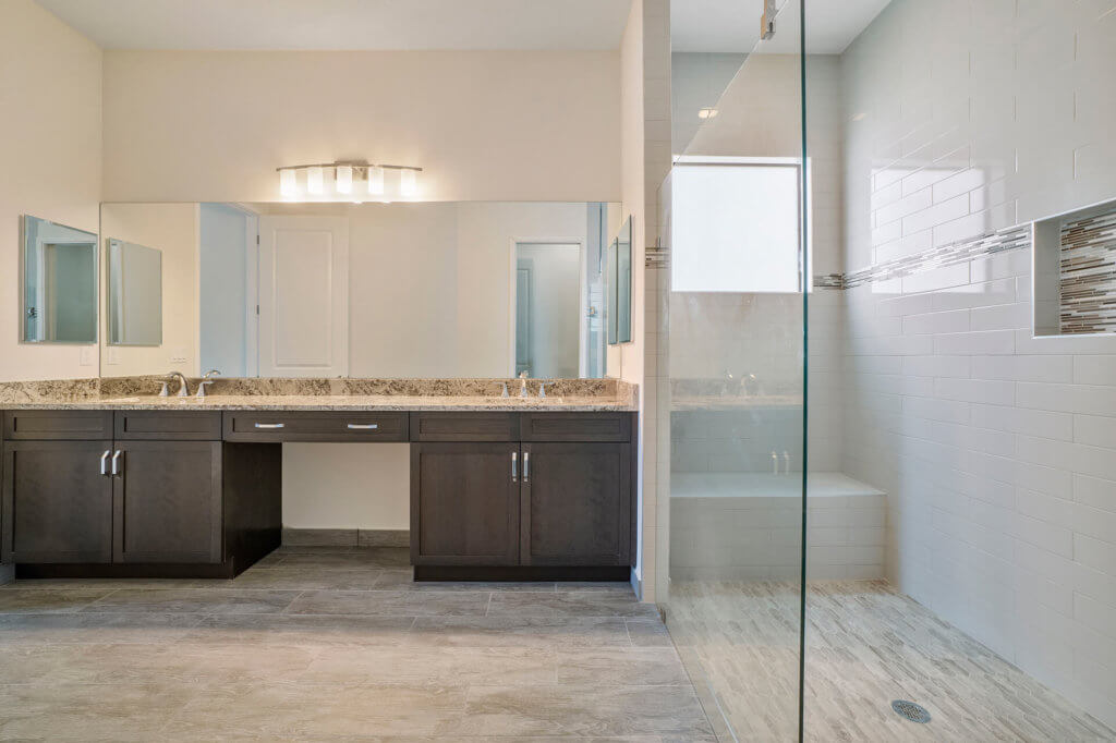 Discover the elegance of a Villamar at Toscana Isles bathroom, showcasing a large mirror above a double sink with dark wood cabinets and granite countertops. A glass-enclosed shower with white tile walls and a niche for toiletries complements the light gray tile flooring beautifully.