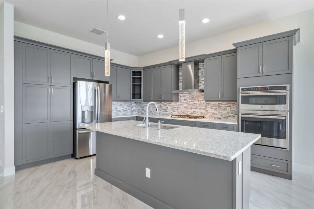 The modern kitchen at Villamar at Toscana Isles boasts gray cabinets, a large central island with a sink, stainless steel appliances, and pendant lighting. The backsplash features a mosaic tile design while light-colored floors elegantly contrast the cabinetry.