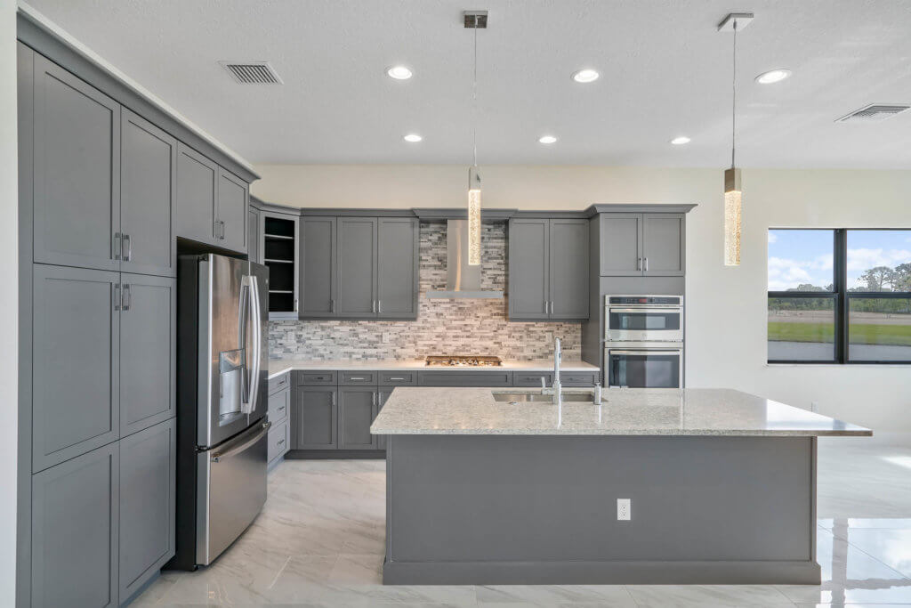 The modern kitchen at Villamar at Toscana Isles boasts gray cabinets, a stainless steel refrigerator, a built-in oven, and a gas stove. A light-colored granite island with a sink sits beneath two pendant lights, while large windows bring in abundant natural light.