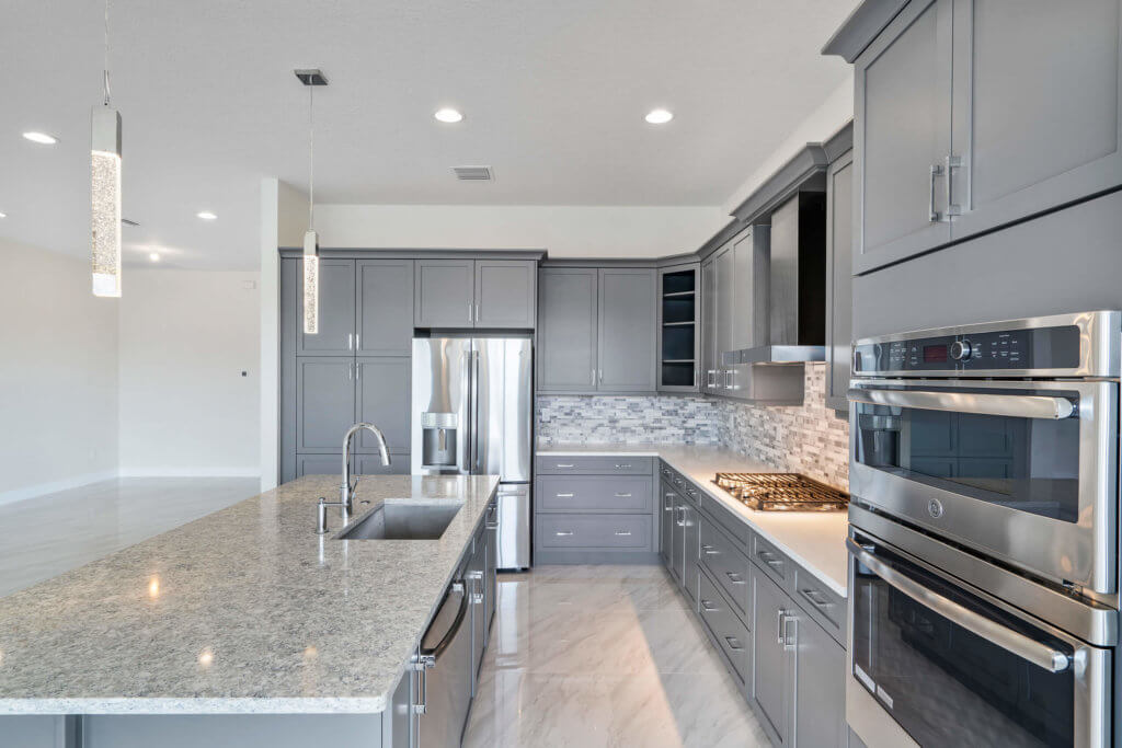 A modern kitchen at Villamar at Toscana Isles boasts gray cabinetry, stainless steel appliances, and a marble-tiled backsplash. The space features a large island with a sink and granite countertop. Pendant lights hang gracefully from the ceiling, complementing the light-colored tiled floor.