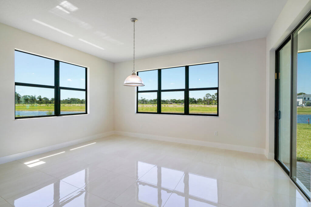 A bright, empty room at Villamar at Toscana Isles features large windows on two walls, a modern pendant light hanging from the ceiling, and glossy tiled floors. The windows offer a view of a grassy field and a partially visible pond outside.