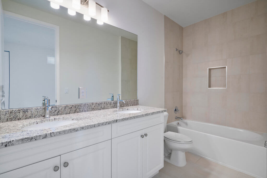 A modern bathroom at Villamar at Toscana Isles features a large mirror above a double sink vanity with granite countertops. The light-colored walls complement the bathtub and shower on the right, while ample ceiling lights ensure the room is perfectly illuminated.