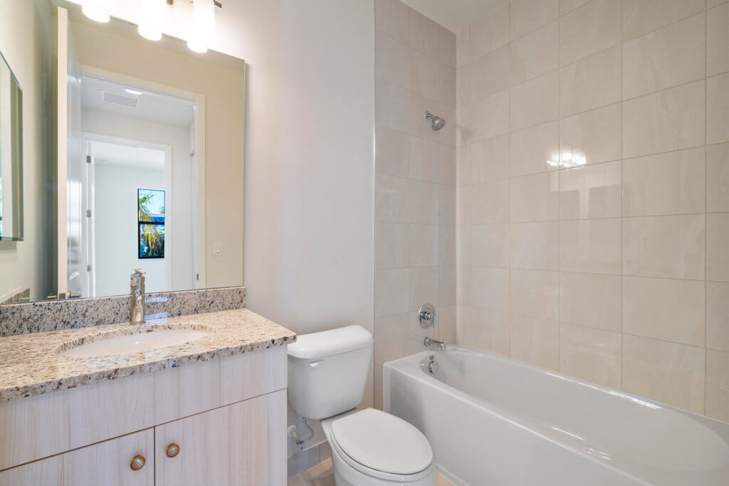 This Villamar at Toscana Isles bathroom features a bathtub and shower, a toilet, and a vanity with a granite countertop and sink. The tiled walls and floors complement the mirrored sink area, while bright overhead lighting ensures the space is always well-lit.