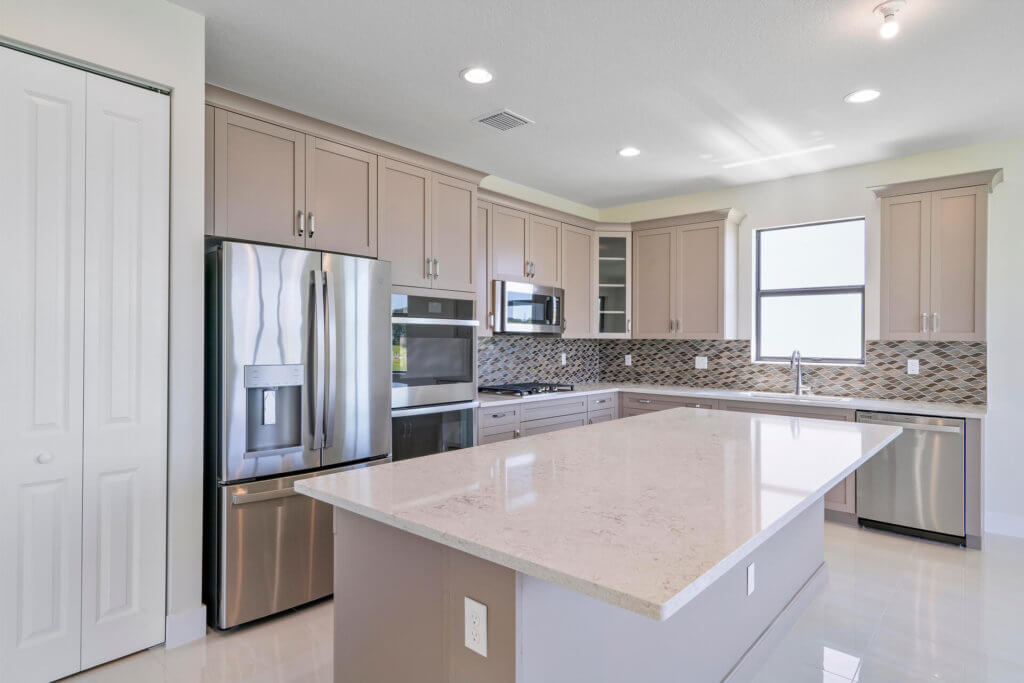 The modern kitchen at Villamar at Toscana Isles features beige cabinets, a stainless steel fridge, and a built-in oven with a gas stove. An island with a light countertop anchors the space, while a window and tiled backsplash add brightness. Light walls and recessed lighting complete the look.