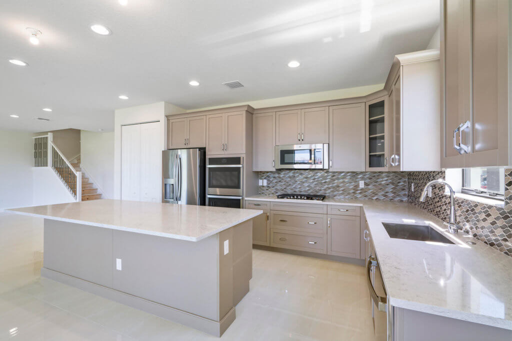 A modern kitchen at Villamar at Toscana Isles boasts light beige cabinetry, a large central island, and stainless steel appliances. The backsplash features a diamond pattern. A staircase is visible in the background, and the room is brightly lit with recessed ceiling lights.