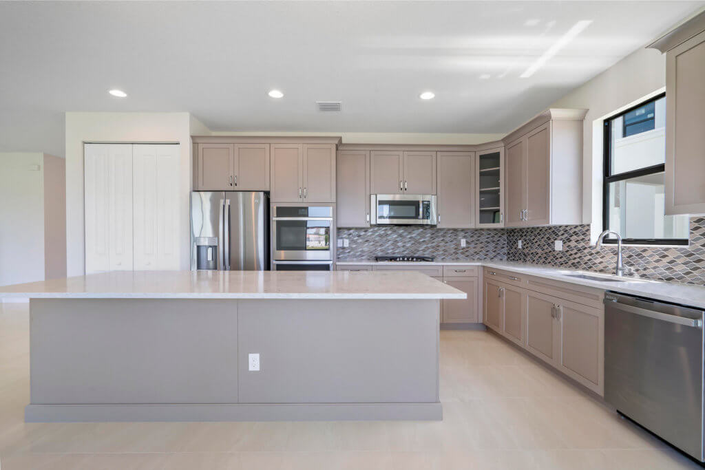 Modern kitchen in Villamar at Toscana Isles with light brown cabinets, stainless steel appliances, and a tiled backsplash. It features a large central island with a marble countertop, microwave, oven, refrigerator, and dishwasher under a bright ceiling with recessed lights.