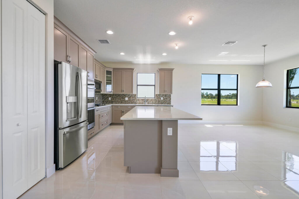 The modern kitchen and dining area at Villamar at Toscana Isles showcases neutral tones, featuring a central island with a light countertop, stainless steel appliances, and beige cabinets. Large windows offer abundant natural light over the glossy tile floor, complemented by an elegant pendant lamp.