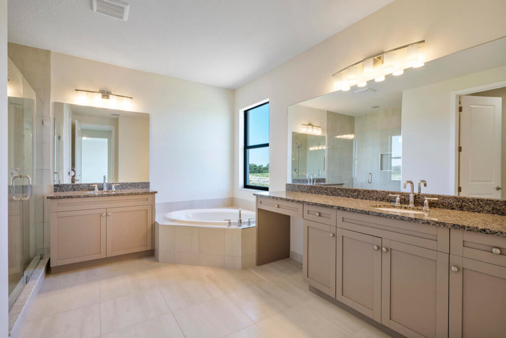 The luxurious Villamar at Toscana Isles offers modern bathrooms featuring a large mirror above a double sink vanity with granite countertops. A corner bathtub is elegantly set against a glass shower enclosure, while ample ceiling lights and natural daylight illuminate the light-tiled floor.