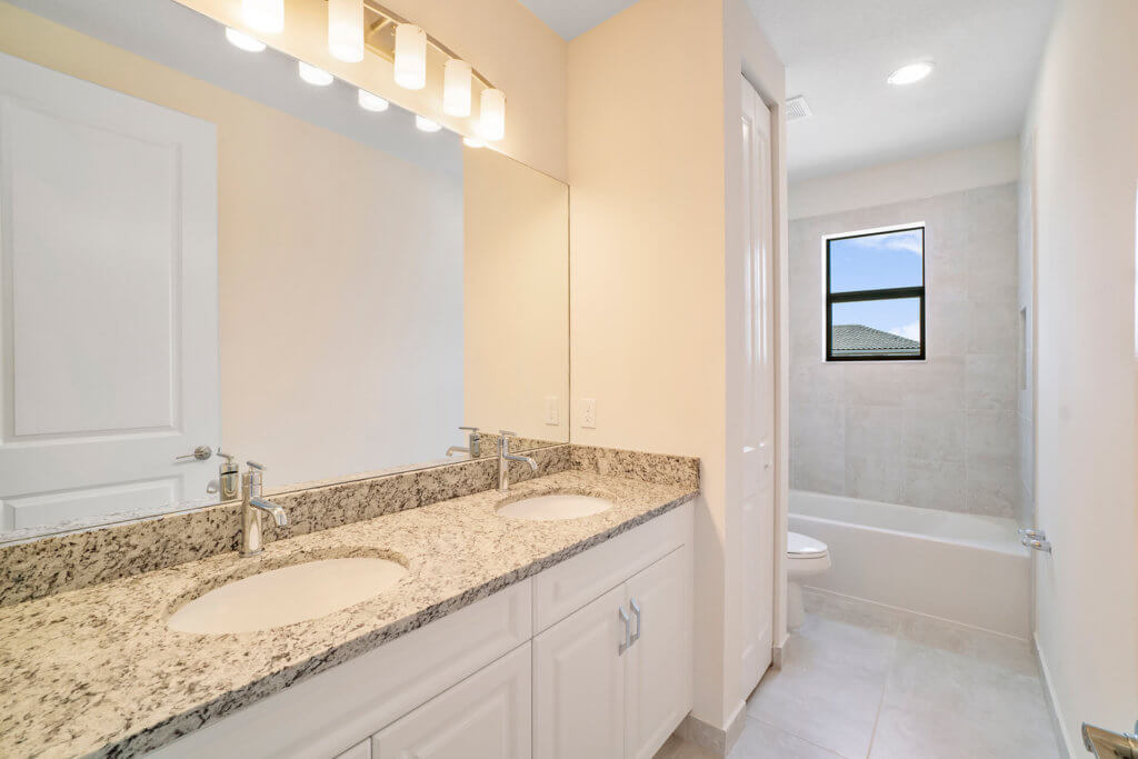 A bathroom at Villamar at Toscana Isles features a granite countertop with two sinks and a large mirror. The room is well-lit with overhead lights. In the background, there is a bathtub with a tiled wall and a small window. The walls are painted light beige.