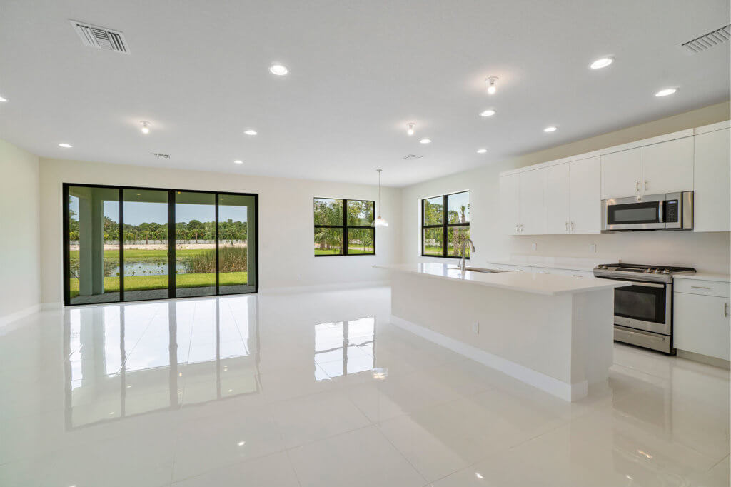 A spacious, modern kitchen and living area at Villamar at Toscana Isles features white glossy floor tiles, sleek cabinetry, and stainless steel appliances. Large windows and glass sliding doors provide a picturesque view of greenery and the river outside. The room is well-lit with recessed lighting.