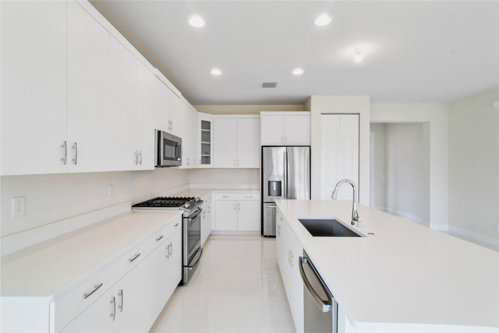 A modern kitchen at Villamar at Toscana Isles features white cabinets, a stainless steel refrigerator, oven, and microwave. The large island boasts a sink and dishwasher. Pristine white countertops complement the recessed lighting tastefully installed in the ceiling.