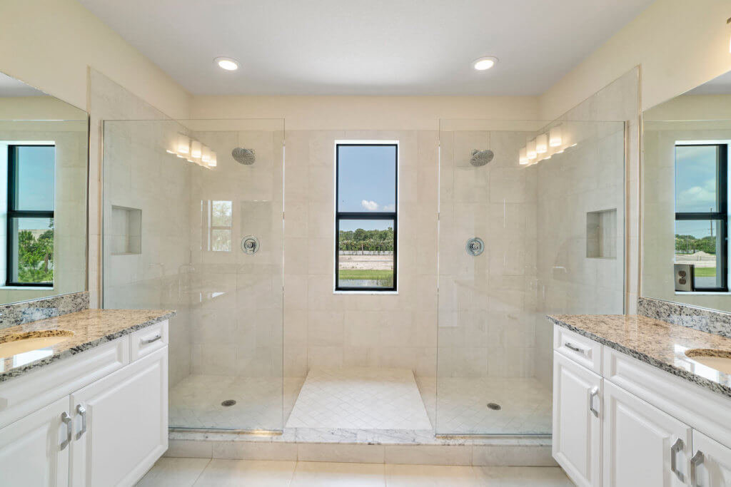 The modern bathroom at Villamar at Toscana Isles features a large double shower with glass doors and two showerheads, centered by a window with an outdoor view. On each side, white cabinets with granite countertops offer style and function. White tile flooring completes the space.
