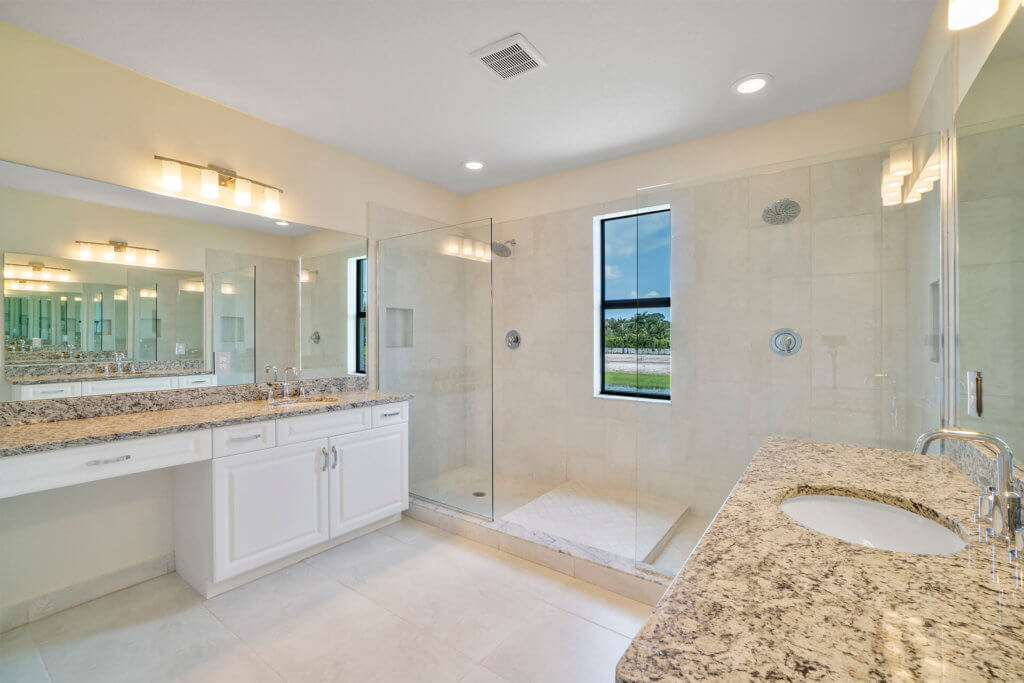 Experience luxury at Villamar at Toscana Isles with a modern bathroom featuring a large glass-enclosed shower, dual sinks on a marble countertop, and spacious white cabinetry. A wide mirror graces the area above the sinks, while a window bathes the space in natural light.