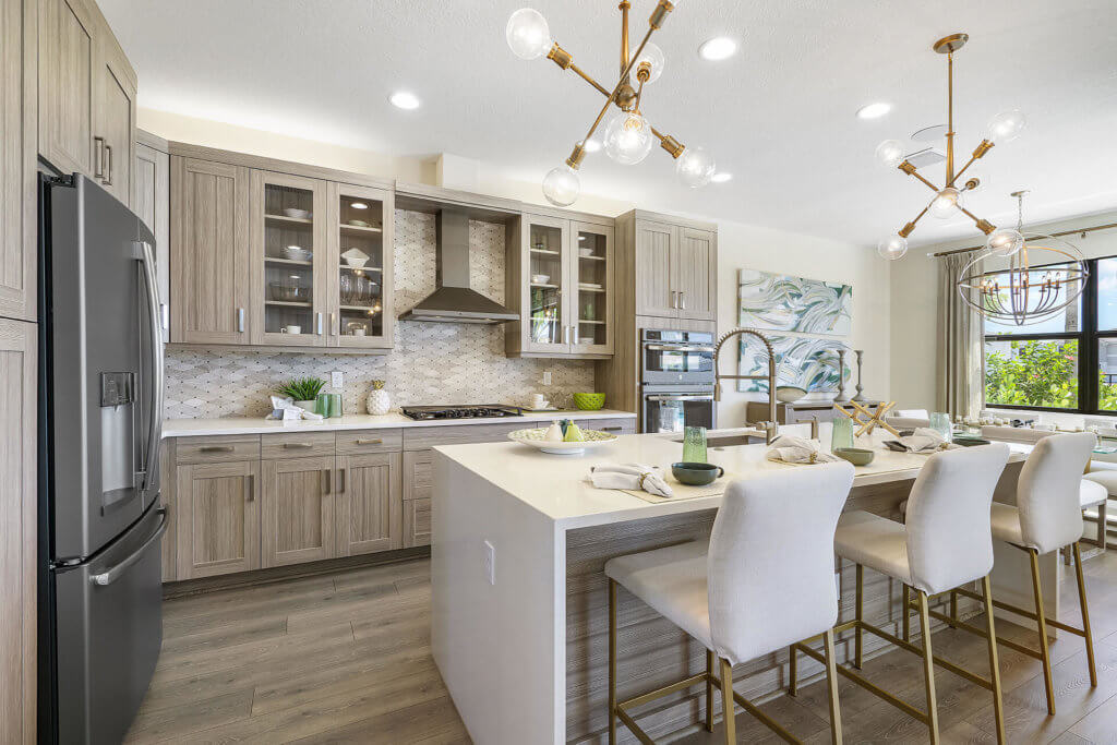 This modern kitchen at Villamar at Toscana Isles boasts light wood cabinets, an island with barstools, and sleek stainless steel appliances. Geometric chandeliers illuminate the space, while a mosaic-patterned backsplash adds flair. A large window welcomes natural light with style.