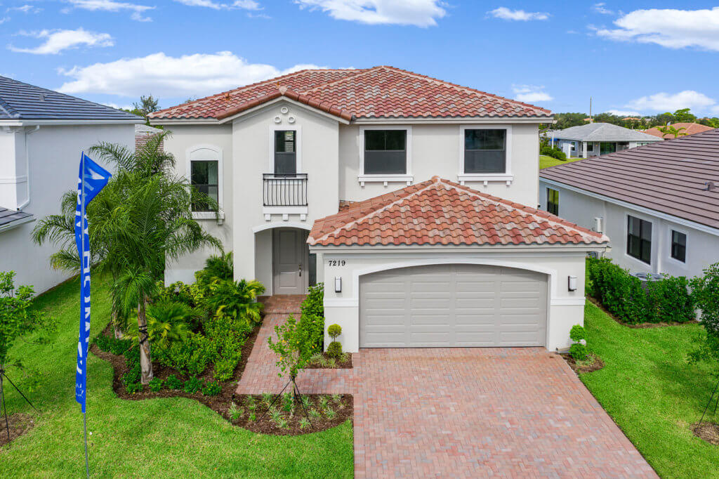 A two-story house with a red-tiled roof and attached two-car garage, nestled in the desirable Villamar at Toscana Isles. The front yard features a paved driveway, landscaped greenery, and a vertical blue sign on the left. Other homes and lush lawns stretch under a partly cloudy sky.