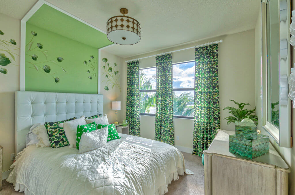 A bright bedroom at Villamar at Toscana Isles features a green accent wall, a white tufted headboard, a bed with green and white pillows, and green leaf-patterned curtains. A ceiling light fixture hangs above, and a dresser with a potted plant stands near the window.
