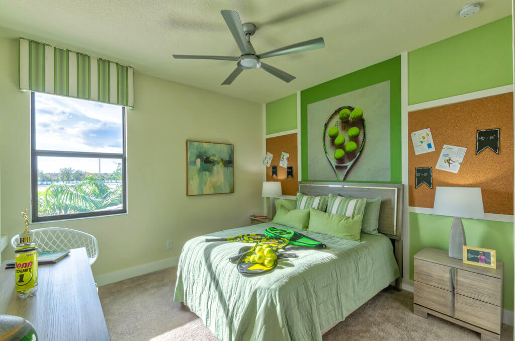 A bright bedroom at Villamar at Toscana Isles features a green and beige color scheme. The bed has green bedding with lemon-themed decorative pillows. A ceiling fan presides over the room, while the window offers a view of trees. A desk and nightstand complete this serene space.