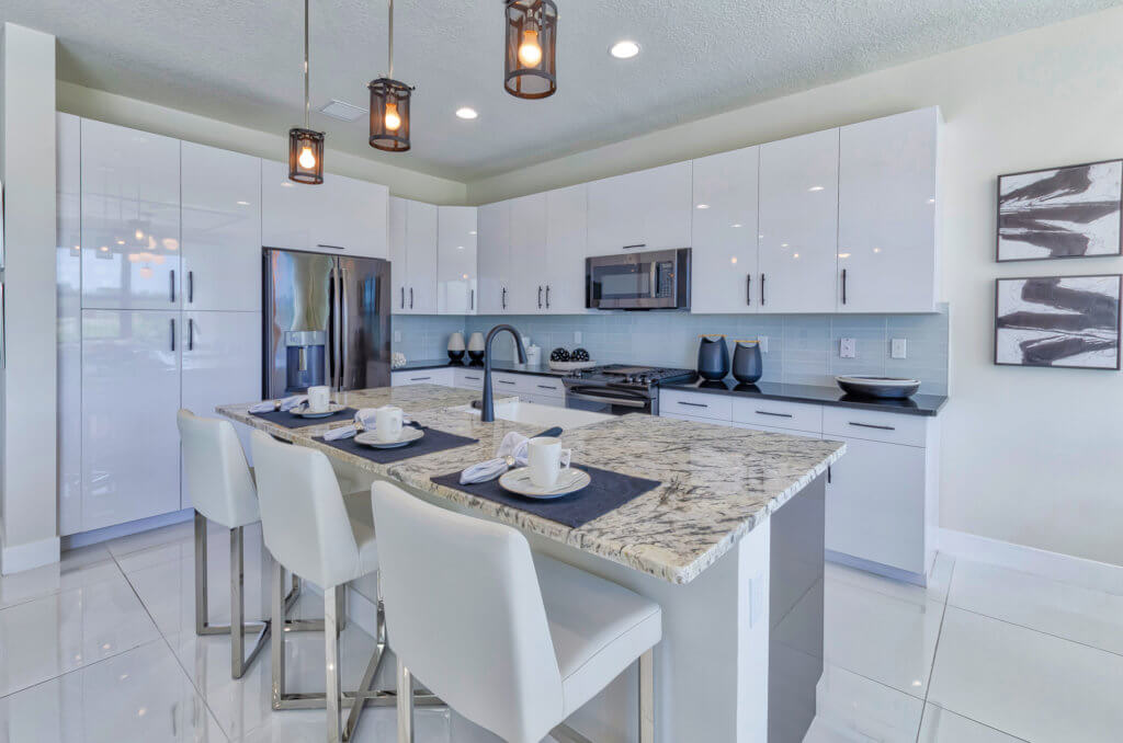 Experience the elegance of a modern kitchen at Villamar at Toscana Isles, boasting white cabinets and a large island with a granite countertop. Three bar stools line the island under stylish pendant lights, while stainless steel appliances complement the spacious tile flooring.