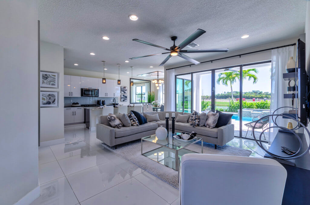 The modern living room at Villamar at Toscana Isles boasts a neutral color palette, featuring two sofas, a glass coffee table, and a ceiling fan. Large sliding glass doors reveal a lush green view. The open kitchen area is equipped with white cabinetry and stainless steel appliances.