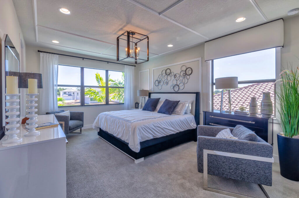 A modern bedroom at Villamar at Toscana Isles features a large bed, gray upholstered chairs, and a dresser. The room boasts light gray walls, a decorative wall piece above the bed, and a large window with views of a tiled roof and greenery outside.