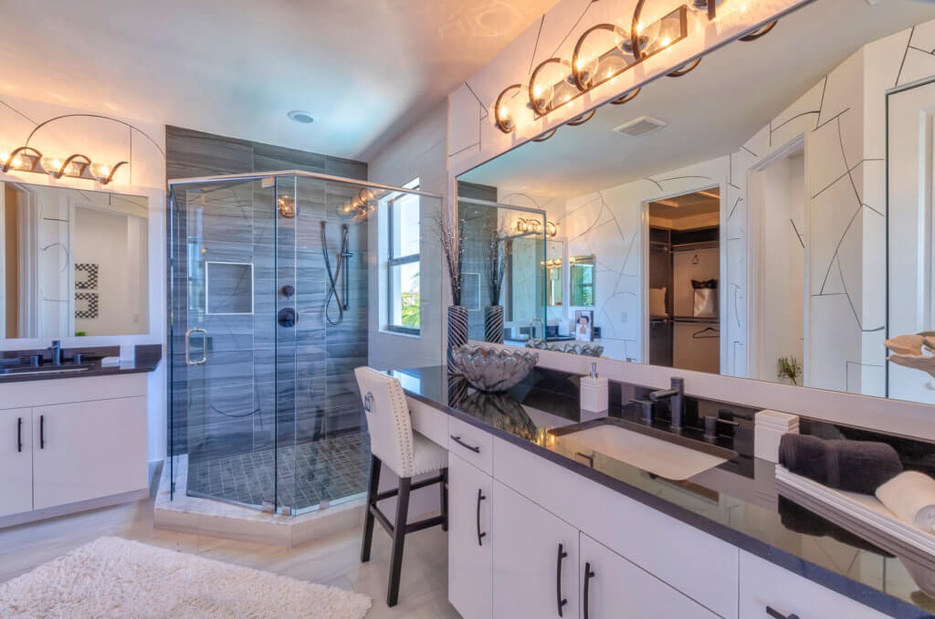 A modern bathroom at Villamar at Toscana Isles features a glass-enclosed shower with gray tiles, a large mirror with overhead lights, a white double-sink vanity with black countertops, and a white cushioned chair. A decorative bowl graces the counter, and a plush white rug adorns the floor.