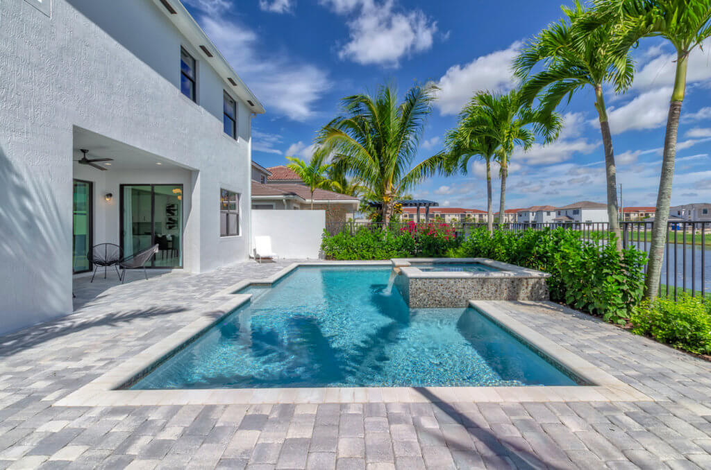 The swimming pool at Villamar at Toscana Isles boasts a built-in hot tub and is framed by a stone patio. The modern house beside it features large glass doors, while tropical plants and palm trees enhance the serene vibe. Above, a blue sky with scattered clouds completes this picturesque setting.