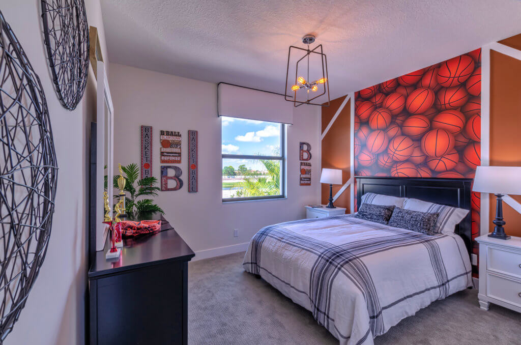 A bedroom at Villamar at Toscana Isles boasts a basketball-themed accent wall, complemented by a bed with plaid bedding. Two lamps flank the bed, while large decorative spheres adorn the left wall. A window offers natural light and a view of tranquil trees outside.