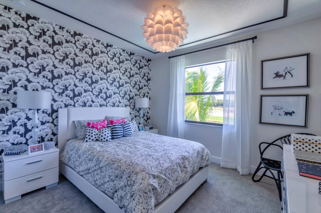 A bedroom at Villamar at Toscana Isles showcases a bed with a floral-patterned duvet and multiple pillows. Behind the bed, black and white patterned wallpaper adds elegance. A large pendant light illuminates the space, which includes a window, side tables, and framed art for added charm.