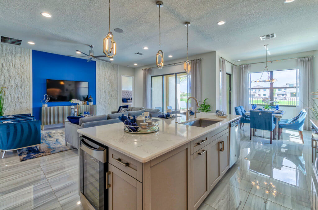 A modern open-concept living room and kitchen at Villamar at Toscana Isles. The kitchen boasts an island with a sink and pendant lighting, while the living area features a large blue TV screen and gray sectional sofa. A dining table with blue chairs sits gracefully near the windows.