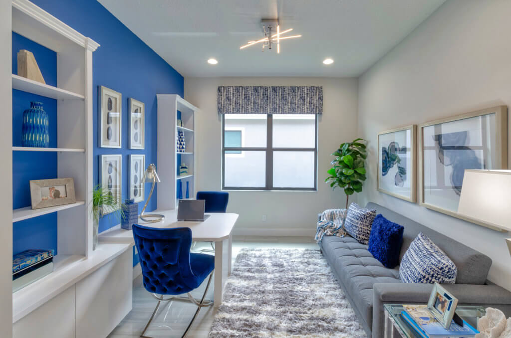 A modern home office in Villamar at Toscana Isles boasts blue and white decor. It features a gray sofa with blue cushions, a white desk with a matching chair, built-in shelves, framed artwork, and a large window with patterned curtains—all brought together by a soft gray rug on the floor.