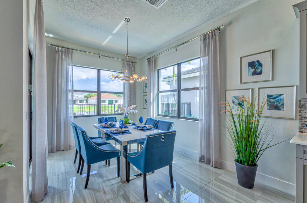 In the modern dining room at Villamar at Toscana Isles, a rectangular table set for six is surrounded by blue upholstered chairs. Large windows bathe the space in natural light, while light curtains, framed artwork, a hanging light fixture, and a tall plant add to the ambiance.