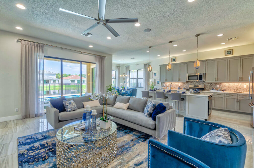 The modern open-concept living area at Villamar at Toscana Isles features a grey sofa, blue armchairs, and a glass coffee table. In the background, the kitchen boasts pendant lights, an island with barstools, and stainless steel appliances. Large windows provide ample natural light.