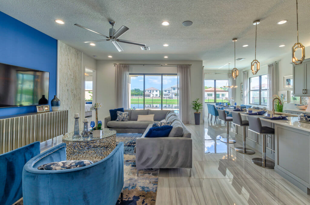 Modern open-concept living room and kitchen at Villamar at Toscana Isles with blue and gray decor. A sectional sofa and chairs face a wall-mounted TV. The kitchen features a counter with barstools and pendant lights, while large windows showcase views of neighboring houses and greenery.