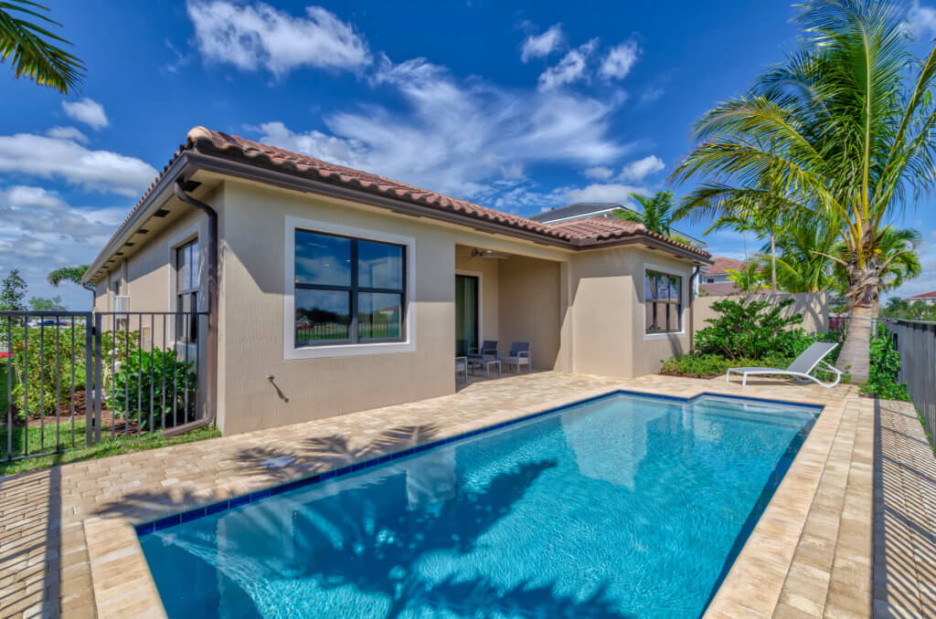 A small backyard at Villamar at Toscana Isles features a rectangular swimming pool bordered by a paved deck. The adjacent beige house boasts large windows and a tiled roof. A few chairs and a palm tree sit next to the pool, with the sky above scattered with clouds.