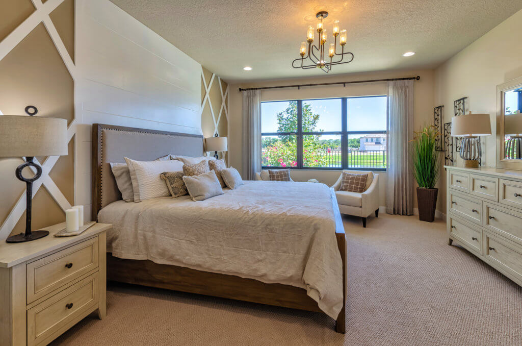 A well-lit bedroom at Villamar at Toscana Isles features a large bed with neutral bedding, flanked by two bedside tables with lamps. The room boasts a patterned accent wall, an armchair, a dresser, and views of lush green trees through the window. A modern chandelier graces the ceiling.