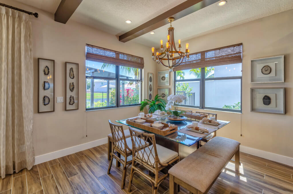 In the dining room of Villamar at Toscana Isles, a wooden table set for a meal is surrounded by chairs and a bench. A chandelier hangs from the ceiling, while large windows bathe the space in natural light. Art adorns the walls, complementing the warm wood finish of the floor.