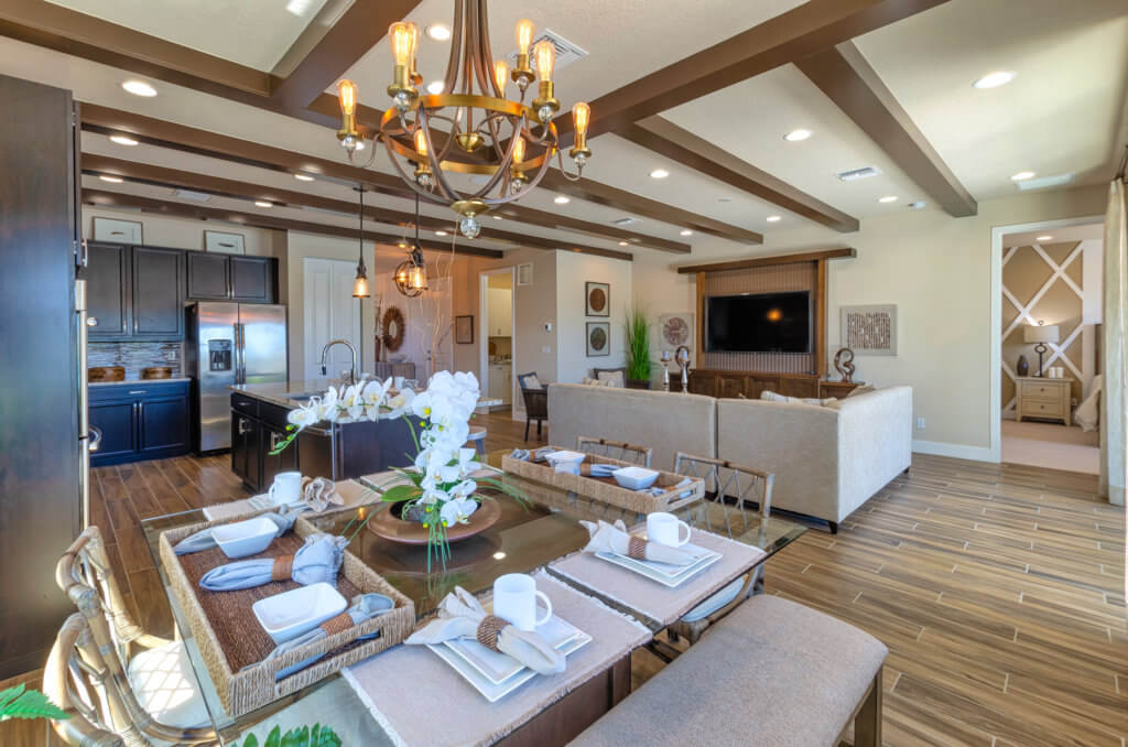 The modern open-concept living and dining area at Villamar at Toscana Isles boasts a wooden table set with plates and napkins. A chandelier elegantly hangs above, while a kitchen with dark cabinets is visible in the background, all adorned in neutral tones and wood accents.