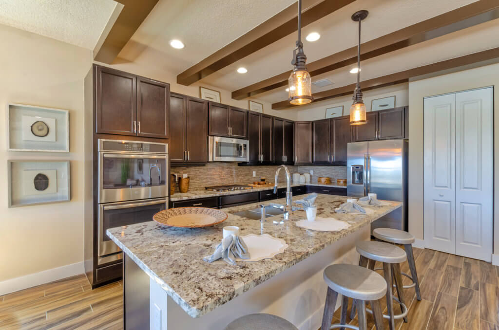 Discover the modern kitchen at Villamar at Toscana Isles, featuring dark wood cabinets, stainless steel appliances, and a large granite-topped island. Three wooden barstools invite you to relax under stylish pendant lights while decorative items and framed artwork add charm to the walls.